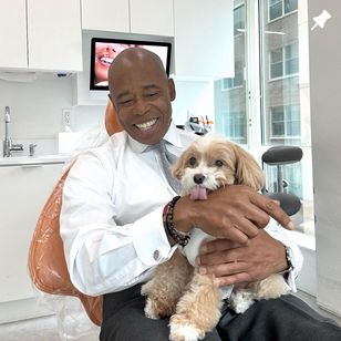 A man in a white shirt sits in a dental office chair, smiling and holding a small dog that has its tongue out.